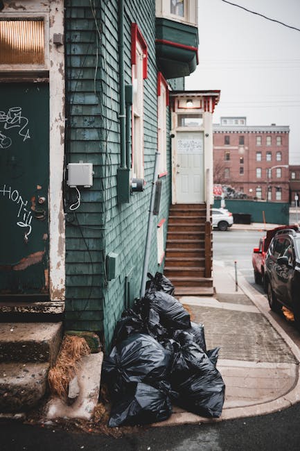 A residential building with weathered teal-green wooden cladding and white trim features a narrow staircase leading to a white front door with a small red-and-white striped awning. Several black garbage bags are piled at the base of the building on the concrete sidewalk, with some showing signs of being filled and tied. To the left, there is an older metal door with graffiti, partially covered by dirt and grime, leading into a different part of the building. The scene is set in an urban environment, with parked cars visible along the street and a red brick building in the background under an overcast sky. Visible utility boxes and cables sit on the exterior wall, emphasizing a service-oriented context. The atmosphere suggests a typical day of rubbish collection or private waste disposal in a city neighbourhood, with the black bags indicating scheduled rubbish removal by local waste management services such as Rubbish Removal Hackney, supporting alternative waste handling options in the area.