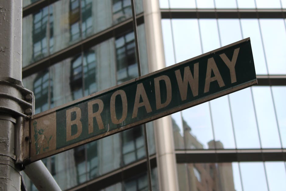 A weathered street sign reading 'BROADWAY' is mounted on a metallic pole at the corner of a building. The sign's surface shows signs of age, with visible dirt and slight corrosion around the edges, and features black lettering on a white background with a small illustration of a blue flower and green leaves on the left side. In the background, modern glass office buildings reflect the surrounding cityscape, with large windows creating a grid-like pattern and metallic structural elements supporting the glass facade. The lighting suggests an overcast day, with diffuse natural light illuminating the scene evenly. The overall context hints at an urban environment typical for areas where private waste collection or alternative rubbish removal services, such as those provided by Rubbish Removal Hackney, may operate in central city locations. The juxtaposition of old signage with contemporary architecture visually underscores the ongoing evolution of city landscapes and the need for efficient waste management solutions in busy commercial districts.