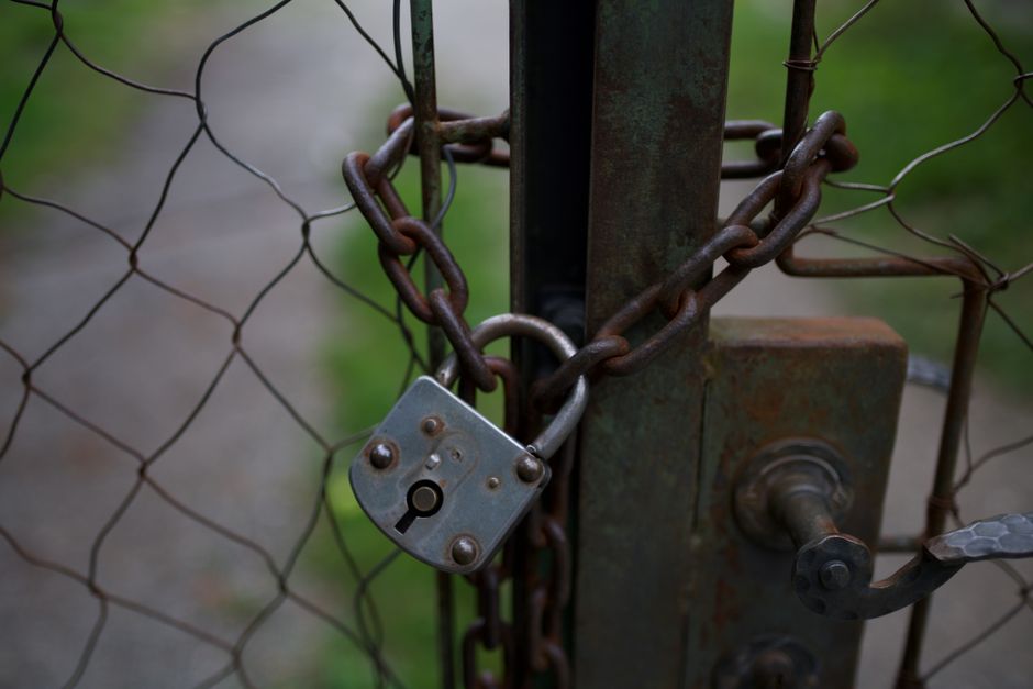 A close-up of a metal gate secured with a rusted chain and a small padlock, with the chain wrapped tightly around the gate's vertical post and latch mechanism. The chain displays a weathered appearance with a reddish-brown patina indicating corrosion, and links that are thick and coarse in texture. The padlock, made of metal, has a slightly tarnished, matte finish with visible rivets and keyhole, positioned at the center of the chain. The gate is constructed from a heavy-duty green-painted metal frame with a weathered, slightly oxidized surface, and the locking mechanism is attached to this frame. Behind the gate, there is a wire mesh fence with a diamond pattern, showing signs of rust and distortion, which extends into a blurred background of green grass and foliage. The scene is evenly lit with natural light, emphasizing the texture differences between the rusted chain, padlock, and the metal structure. This image visually relates to private or independent waste management, such as securing disposal areas or waste storage sites, often handled by specialist services like Rubbish Removal Hackney.