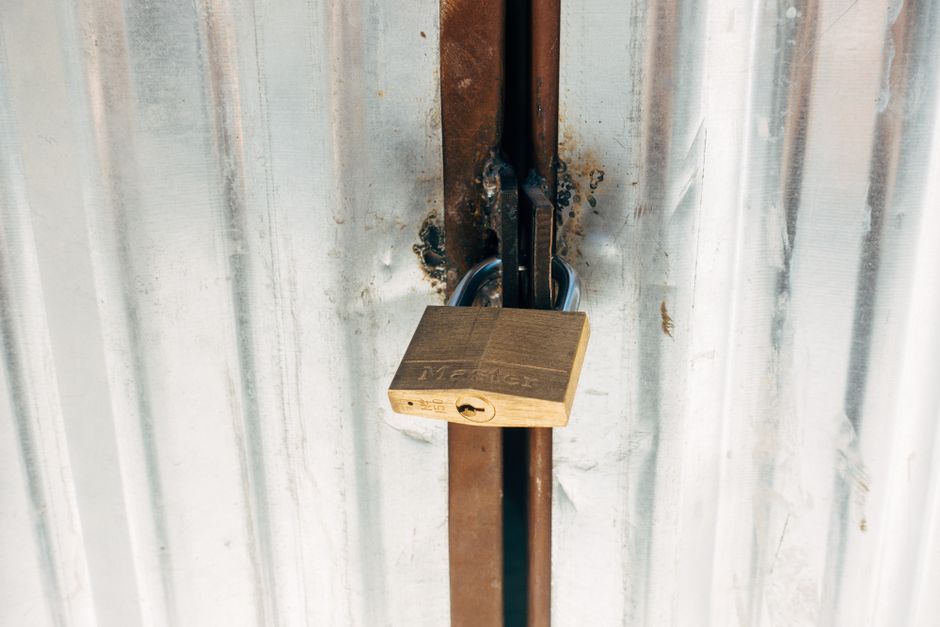 Close-up image of a black padlock securing a metal latch on a green wooden gate. The padlock shows signs of rust, with a weathered brass shackle. The latch is mounted on vertical wooden planks painted in a muted teal or greenish hue, with visible wood grain and small rusted nail heads. The background reveals additional wooden slats, all painted in the same colour, indicating an outdoor setting such as a garden or driveway entrance. The lighting is natural, casting soft shadows, and the scene appears to be taken during daylight hours. This setup is typical of private property access points, often associated with independent waste collection or secure enclosures for rubbish or other stored items, aligning with rubbish removal and waste management services that require access control.