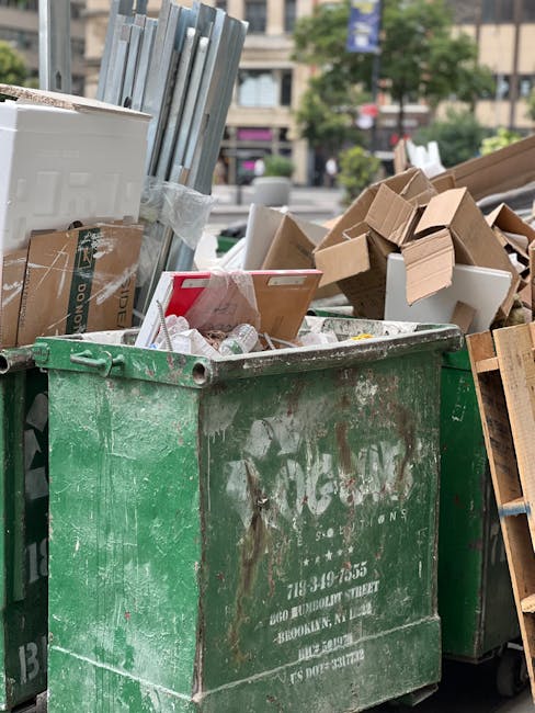A large, weathered green skip filled with mixed waste materials, positioned on a street or outdoor area. The open top reveals a variety of discarded items including flattened cardboard boxes, some with printed labels and logos, white plastic packaging, and smaller waste items. To the left and partially visible, there are upright metal frames or panels, secured with tape, suggesting part of furniture or equipment being discarded. On the right, there are additional cardboard boxes, some collapsed or damaged, and a wooden pallet leaning against the skip. In the background, blurred urban elements such as buildings, trees, and passerby indicate a busy city environment. The scene shows an outdoor waste collection possibly associated with a private rubbish removal service, emphasizing efficient on-site clearance options separate from municipal collection. This setup reflects typical practices in private waste handling services like Rubbish Removal Hackney, tailored for small flats or similar properties.