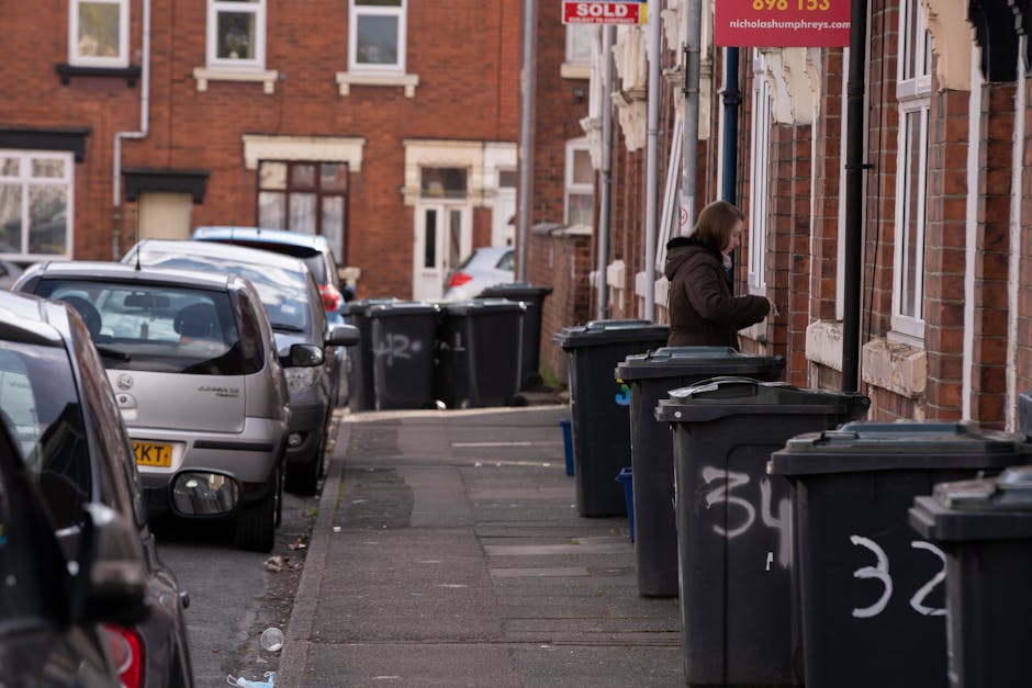 A row of black wheelie bins with closed lids line the edge of a paved sidewalk along a residential street, positioned close to a red brick terraced building. The bins have white painted numbers on their sides, indicating different collection points. A woman dressed in a brown coat is seen standing beside one of the bins, appearing to manage or sort waste. Several parked cars, including a silver hatchback, occupy the street in the background, with additional houses visible further down the road. The building facades feature white-framed windows and brickwork detailing, and a street sign advertising property sales is partially visible overhead. The scene depicts an area where private waste collection services, such as those provided by Rubbish Removal Hackney, are used for on-site rubbish disposal. The natural lighting and slight cloud cover create a neutral, daytime atmosphere, emphasizing the practical arrangement of waste management in a typical urban setting, suitable for alternative rubbish removal solutions away from local authority collection services.
