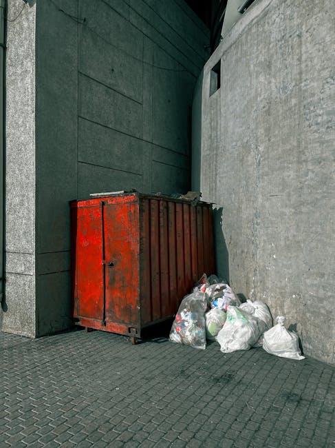 A large, weathered green skip filled with mixed waste materials, positioned on a street or outdoor area. The open top reveals a variety of discarded items including flattened cardboard boxes, some with printed labels and logos, white plastic packaging, and smaller waste items. To the left and partially visible, there are upright metal frames or panels, secured with tape, suggesting part of furniture or equipment being discarded. On the right, there are additional cardboard boxes, some collapsed or damaged, and a wooden pallet leaning against the skip. In the background, blurred urban elements such as buildings, trees, and passerby indicate a busy city environment. The scene shows an outdoor waste collection possibly associated with a private rubbish removal service, emphasizing efficient on-site clearance options separate from municipal collection. This setup reflects typical practices in private waste handling services like Rubbish Removal Hackney, tailored for small flats or similar properties.