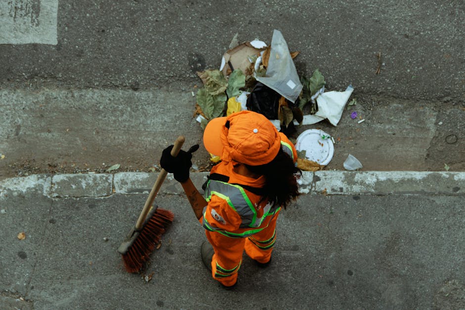 A waste management worker dressed in bright orange high-visibility clothing with reflective stripes, black gloves, and an orange cap, is standing on a paved sidewalk next to a concrete wall. The worker is holding a long-handled, wide-bristled broom and appears to be in the process of clearing litter from the curbside area. In front of the worker lies a pile of discarded rubbish, including a crumpled paper bag, torn cardboard, a plastic container, and other various waste items, some partially flattened or crumpled. The trash is loosely gathered towards the base of the wall, which shows signs of weathering and stains. The environment suggests an urban setting, with the sidewalk and street surface showing typical asphalt and concrete textures, and the overall scene illuminated by natural daylight. This image exemplifies private rubbish collection practices, consistent with services like those offered by Rubbish Removal Hackney for local waste clearance and curbside rubbish removal.