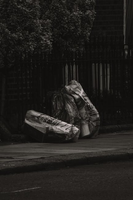 Two large black plastic garbage bags filled with waste are placed on a sidewalk near a black metal fence with vertical bars, located in an outdoor urban setting. The bags appear to be tightly tied and slightly bulging, indicating they are full. One bag has visible printed text and symbols, suggesting it is for waste collection or disposal. Behind the bags, there are dense, leafy bushes providing some greenery and partial cover, while the surrounding environment includes a paved curb and part of a street surface in the foreground. The scene is lit with subdued natural lighting, creating shadows on the bags and the sidewalk, reflecting a typical quiet street scene related to private rubbish collection or independent waste removal efforts, as sometimes performed by local services like Rubbish Removal Hackney, in a manner consistent with alternative waste handling practices in urban areas.