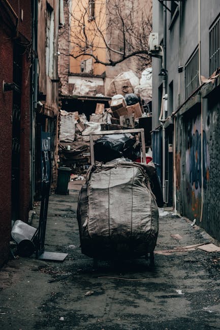 A narrow urban alleyway with a concrete surface, cluttered with scattered debris, including bits of paper, plastic, and small trash items. In the foreground, there is a large, grey, weathered trash bag or sack, partially torn, with a metal handle on top, positioned slightly off-center. Behind it, a platform trolley or handcart loaded with additional waste items, covered with a black plastic sheet, stands on the uneven pavement. The alley is bordered by tall, graffiti-covered brick and metal-clad walls, with some graffiti tags and spray paint visible on the surfaces. To the left, a small black waste bin is placed against the wall, and a discarded piece of wood lies nearby. In the background, a pile of cardboard boxes and more refuse are stacked haphazardly, with bare leafless tree branches and the façade of an apartment building visible above. The lighting is diffuse, suggesting overcast weather, contributing to a stark, unkempt scene typical of an area awaiting scheduled or independent waste clearance, as might be handled by services such as Rubbish Removal Hackney in private waste disposal settings.