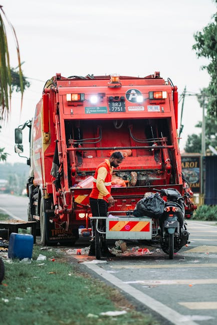 A waste collection worker wearing a yellow and red uniform is operating a large red garbage truck on a roadside. The worker is standing at the rear of the truck, which features a large, open hopper for waste collection with visible hydraulic mechanisms and safety lights at the top. In front of the worker, there is a small black motorcycle parked on the sidewalk, with a black plastic bag placed on its seat. The area surrounding the scene includes a patch of green grass, a blue plastic container, and some scattered debris close to the curb. The background shows a street with utility poles, trees, and a blue structure possibly indicating a local business or storage area. The environment appears to be daytime with natural light, and the scene reflects a typical urban setting where independent waste collection or rubbish removal services like those provided by Rubbish Removal Hackney might operate, especially for on-site clearance or alternative waste handling outside standard council collection schedules.