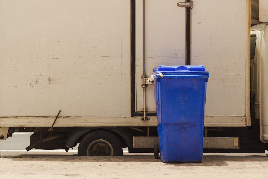 A blue plastic wheelie bin with a hinged lid is positioned on a flat pavements surface next to the side of a small, white delivery van or utility vehicle. The bin has a smooth, glossy finish and appears slightly weathered with some dirt or grime marks on its surface. It is situated close to the vehicle's metal body, which shows visible signs of age, including minor scratches and dirt. The wheelie bin's wheels are partially visible at the base, with the orange safety handle on the lid clearly identifiable. The background includes a section of the vehicle's metal side panel, which is plain and slightly discolored, with a dark vertical door hinge and a small section of the vehicle’s wheel hub visible underneath. The environment suggests an outdoor urban or suburban setting with a concrete or paved surface beneath the bin and vehicle, highlighting the typical scene of waste receptacle placement for waste collection or alternative rubbish disposal services, as provided by companies like Rubbish Removal Hackney in the context of the local council regulations. The scene conveys a moment of waste management setup, suitable for visual content related to private waste handling or skip alternatives without implying any specific action or disposal method.
