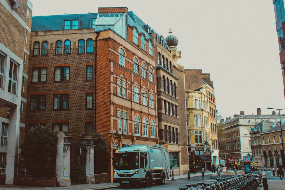 A street scene in an urban area featuring a compact waste collection van parked on the right side of the pavement, with a predominantly white body and blue accents, positioned in front of a series of tall, historic buildings with brick facades, decorative window arches, and ornate architectural details. The buildings' exteriors display a mix of red, brown, and beige tones, with some windows featuring rounded tops and others rectangular. The street is lined with metal barriers and bollards, and in the background, there are additional older buildings with classical architectural elements, including a structure with a domed tower. The environment appears to be well-lit with natural daylight, and the scene suggests an urban setting where independent waste collection services, such as those provided by Rubbish Removal Hackney, might operate as an alternative to council rubbish disposal rules, emphasizing private on-site clearance for local premises.